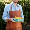 A man in a Personalised Brown Leather Apron and denim shirt holds a green dish with grilled corn cobs while standing outdoors in a garden.