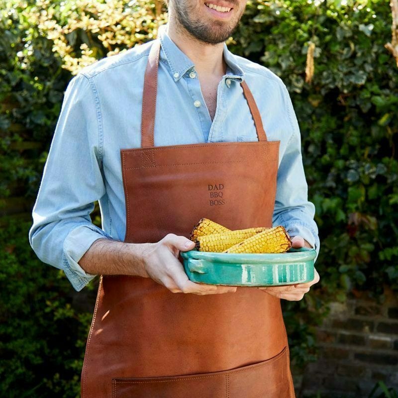 A man in a Personalised Brown Leather Apron and denim shirt holds a green dish with grilled corn cobs while standing outdoors in a garden.