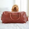 A Personalised Brown Leather Duffle Bag with handles sits on a bed, ideal for travel, while a curly-haired dog relaxes in the background.
