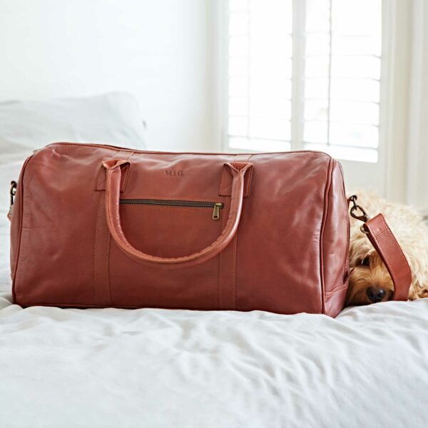 A Personalised Brown Leather Duffle Bag sits on a bed, with a dog partially visible behind it near a window with white blinds.