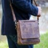A person in a dark jacket and jeans carries a Personalised Brown Buffalo Leather Portrait Messenger Bag with an adjustable shoulder strap outdoors.