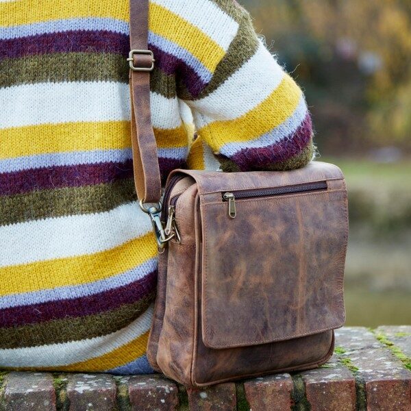 A person in a striped sweater sits on a brick ledge with the Personalised Brown Buffalo Leather Portrait Messenger Bag resting at their side.
