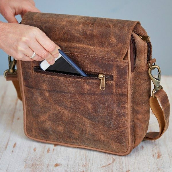 A hand places a smartphone into the front zippered pocket of a Personalised Brown Buffalo Leather Portrait Messenger Bag on a wooden surface.