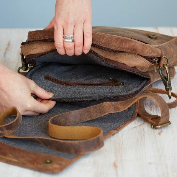 A person opens a Personalised Brown Buffalo Leather Portrait Messenger Bag with a grey fabric lining and multiple zippered compartments on a light wooden surface.