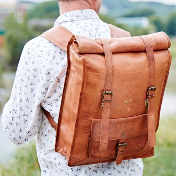 Outdoors, a person in a white patterned shirt carries the Personalised Brown Leather Rolltop Backpack, featuring a large size, buckles, and a front pocket.