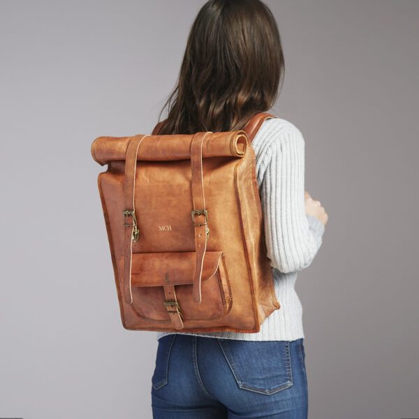 A person in a light sweater and jeans carries the Personalised Brown Leather Rolltop Backpack, featuring buckle closures.