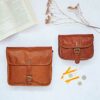 A Personalised Brown Leather Crossbody Bag is placed alongside another bag on the surface, surrounded by an open book, dried flowers, train tickets, and coins.