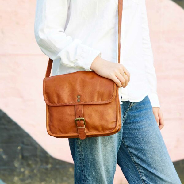 A person wearing a white shirt and blue jeans is holding a Personalised Brown Leather Crossbody Bag with a buckle, standing against a pink and black background.