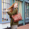 A person with long red hair in a green sweater and gray pants stands outside a building, smiling over their shoulder while holding a Personalised Brown Leather Tote Handbag.