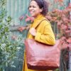 A woman in a yellow coat stands outdoors, smiling, with autumn foliage behind her and carrying a Personalised Brown Leather Tote Shopping Bag on her shoulder.
