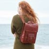 A person with long red hair stands by the sea, wearing a green sweater and carrying a handmade, eco-friendly Personalised Brown Leather Satchel Style Rucksack.