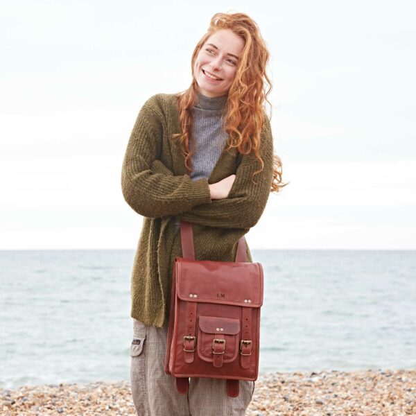 A person with long red hair stands on a pebbled beach, wearing a green eco-friendly sweater and carrying a Personalised Brown Leather Satchel Style Rucksack, with the sea visible in the background.