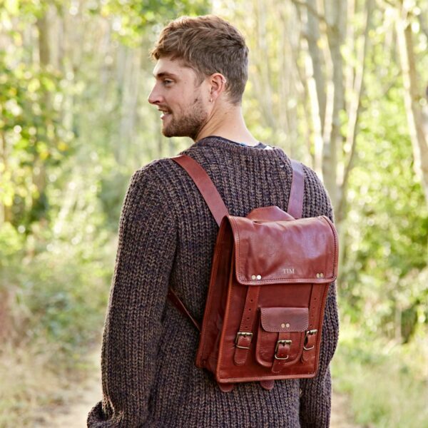 A man in a brown sweater stands outdoors on a forest path, wearing a Personalised Brown Leather Satchel Style Rucksack and looking over his shoulder.