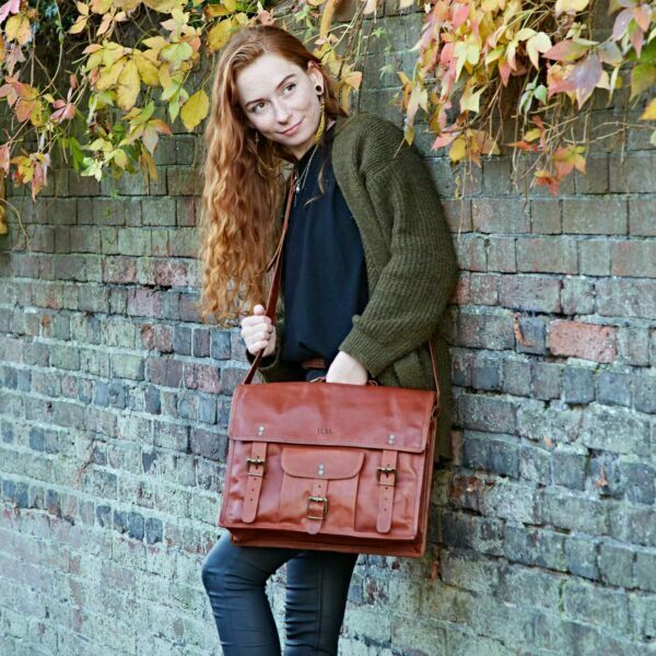 A woman with long red hair stands against a brick wall with autumn leaves, wearing an eco-friendly green sweater and holding a Personalised Brown Leather Vintage Satchel.