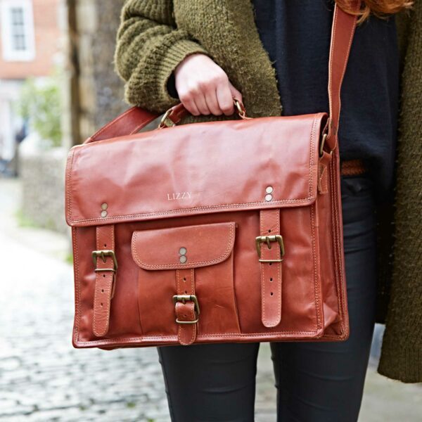 A person holds a Personalised Brown Leather Vintage Satchel with buckles and a top handle, standing on a cobblestone street.