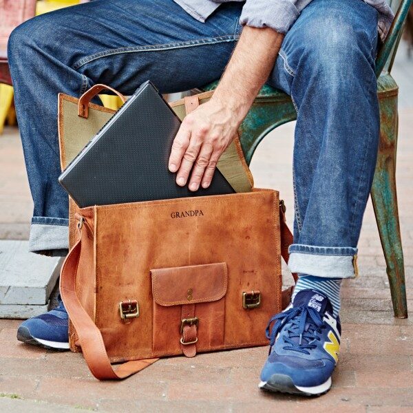 A person in jeans and blue sneakers puts a black laptop into a Personalised Brown Leather Vintage Satchel while sitting on a metal chair.