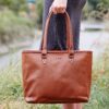 A person in a grey dress holds a Personalised Brown Leather Zipped Tote Shopping Bag with "L.W.B" embossed on the front. Grass and water can be seen in the background.