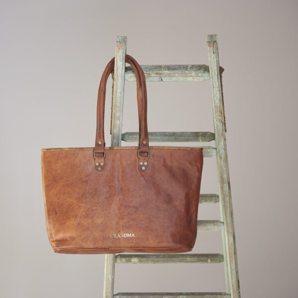 A Personalised Brown Leather Zipped Tote Shopping Bag with "GRANDMA" printed on it hangs from a distressed wooden ladder against a plain background.