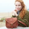 A person with long red hair sits at an outdoor table, with a Personalised Brown Leather Curved Saddle Bag placed in front of them.