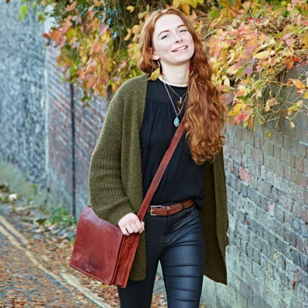A woman with long red hair, wearing a dark jumper and leather trousers, walks outside along a brick wall with autumn leaves, carrying a personalised leather messenger bag.