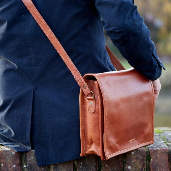 Person in a navy blazer sits on a brick ledge with the Personalised Brown Leather Messenger Bag over their shoulder, a sustainable, handmade accessory featuring 