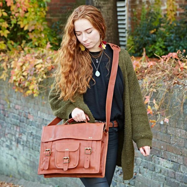 A woman with long red hair walks outdoors, wearing a green cardigan and black shirt, carrying a large brown personalised leather satchel with buckle details.