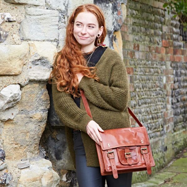 A woman with long red hair leans against a stone wall, wearing a green jumper and carrying a personalised brown leather satchel.