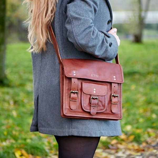 A person outdoors on grass with fallen leaves carries a Personalised Brown Leather Satchel with buckles and the initials 