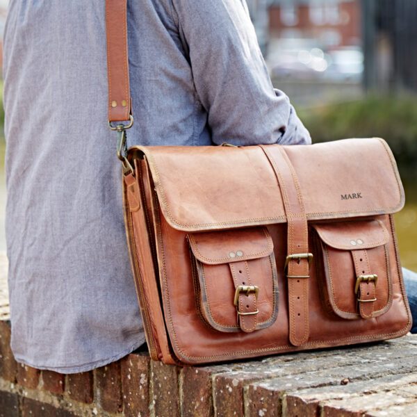 A person wearing a light blue shirt sits on a brick ledge with the Personalised Brown Leather Satchel Bag, which features two front pockets and a shoulder strap.