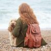 A person with long red hair wearing a sustainable green sweater and carrying a Personalised Large Brown Leather Rucksack sits on a pebbled beach beside a cream-colored dog, facing the sea.