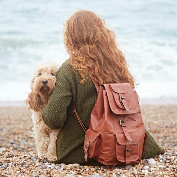 A person with long red hair wearing a sustainable green sweater and carrying a Personalised Large Brown Leather Rucksack sits on a pebbled beach beside a cream-colored dog, facing the sea.