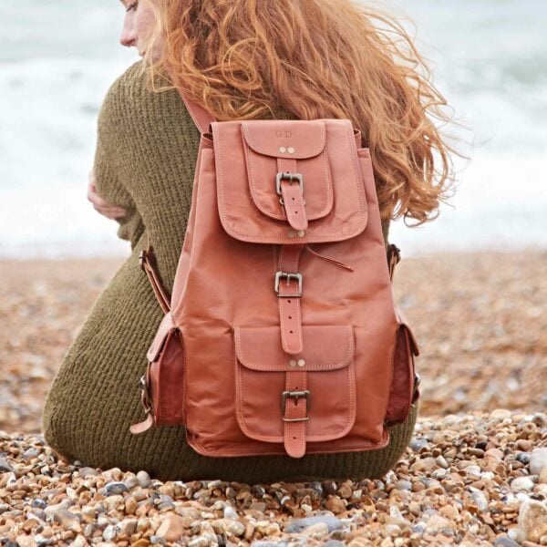 A person with long red hair sits on a rocky beach, facing the water. They wear a green sweater and carry a Personalised Large Brown Leather Rucksack, creating an effortlessly sustainable look.