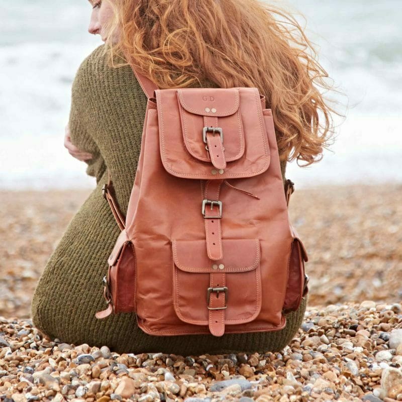 A person with long red hair sits on a rocky beach, facing the water. They wear a green sweater and carry a Personalised Large Brown Leather Rucksack, creating an effortlessly sustainable look.