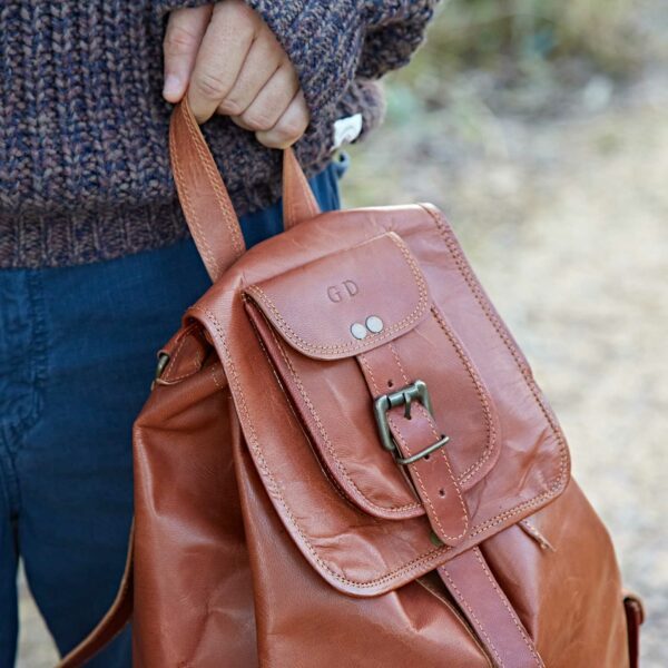 A person holds the Personalised Large Brown Leather Rucksack by its top handle; the front flap, secured with a buckle, is stamped with initials "GD.