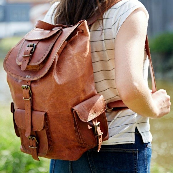 A person outdoors wearing a striped shirt and jeans carries the Personalised Large Brown Leather Rucksack, featuring buckle closures and a handcrafted design.