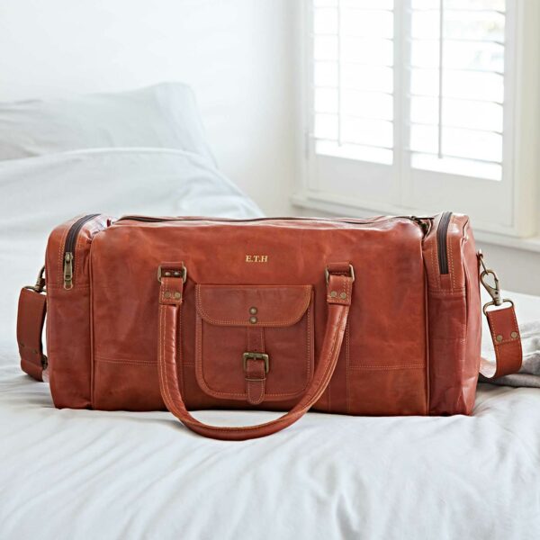 A Personalized Brown Leather Holdall with Zipped Pockets sitting on top of a bed.