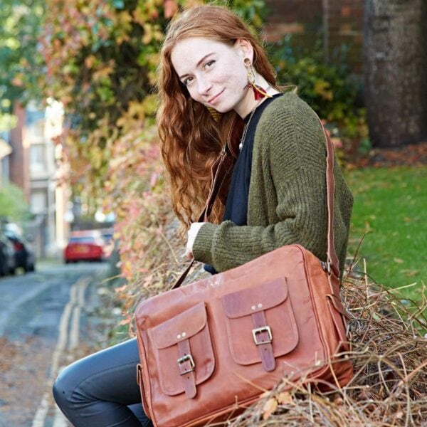 A young woman with long red hair sits outdoors on a low wall, wearing an olive sweater and holding a Personalised Brown Leather Satchel Style Courier Bag.