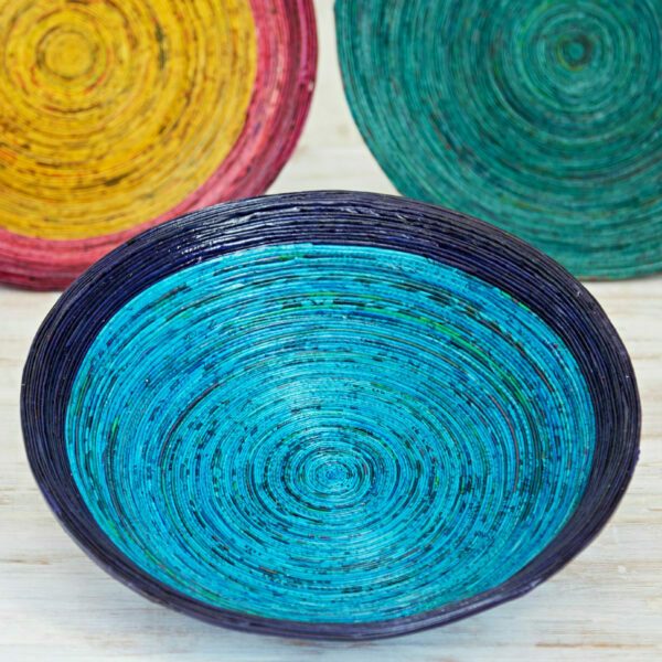 Three colourful, circular woven baskets are displayed on a light wooden surface. The foreground features a striking blue decorative bowl, while the other two in the background are yellow and green.