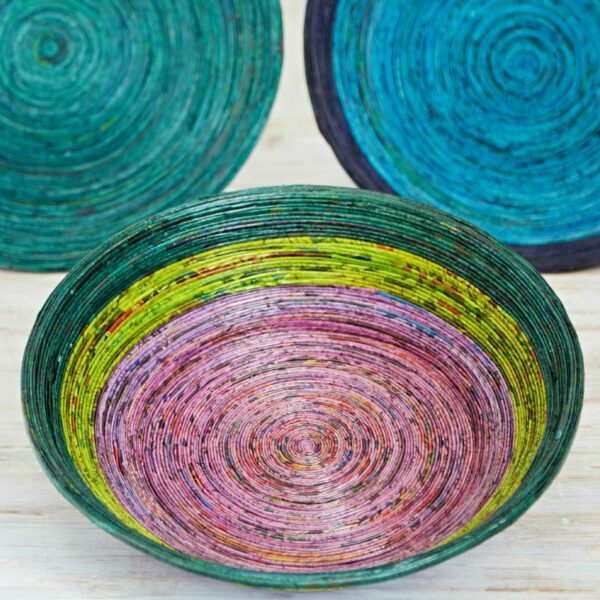 Three colourful, circular coiled baskets made from rolled paper strips are arranged on a light wooden surface. The foreground Recycled Newspaper Bowl features green, yellow, and pink concentric rings—perfect for eco-friendly decor.