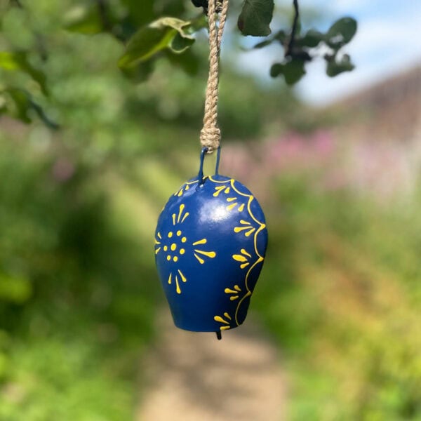 A Hand Painted Blue Iron Cow Bell, featuring beautiful blue and yellow designs, dangles on a rope in an outdoor garden setting with blurred greenery in the background.
