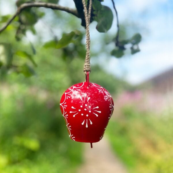 A hand-painted, red, iron cow bell adorned with white floral patterns hangs from a branch outdoors.