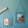 A hand is adjusting a Dia Silver framed artwork with a face drawing and flowers on a blue wall. Next to it is another Dia Silver Recycled Glass Hanging Photo Frame featuring a couple walking on a wooden boardwalk near a body of water.