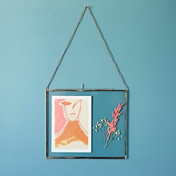 A Dia Silver Recycled Glass Hanging Photo Frame on a blue wall contains an abstract art print and small dried flowers.