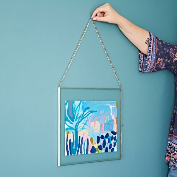 A person carefully hangs a framed abstract painting with a blue and pink color palette on a teal wall, using sleek recycled glass accents to add an eco-friendly touch.