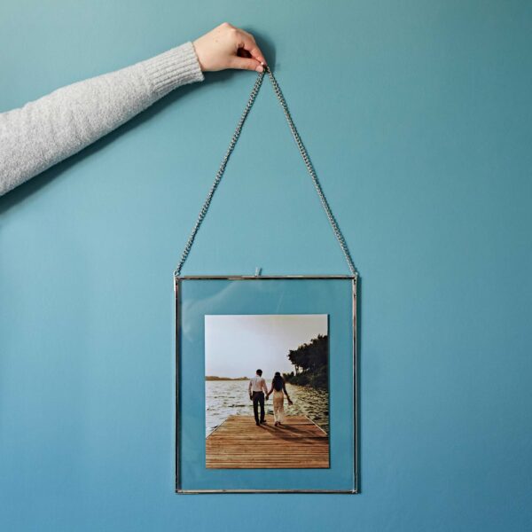 A hand holding a Dia Silver Recycled Glass Hanging Photo Frame with a picture of a couple walking on a wooden pier by the water, set against a blue wall.
