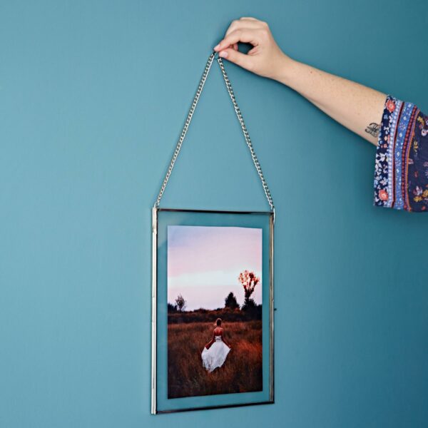 A person carefully hangs a Dia Silver framed photo of a woman in a field on a teal wall, its border crafted from elegant recycled glass.
