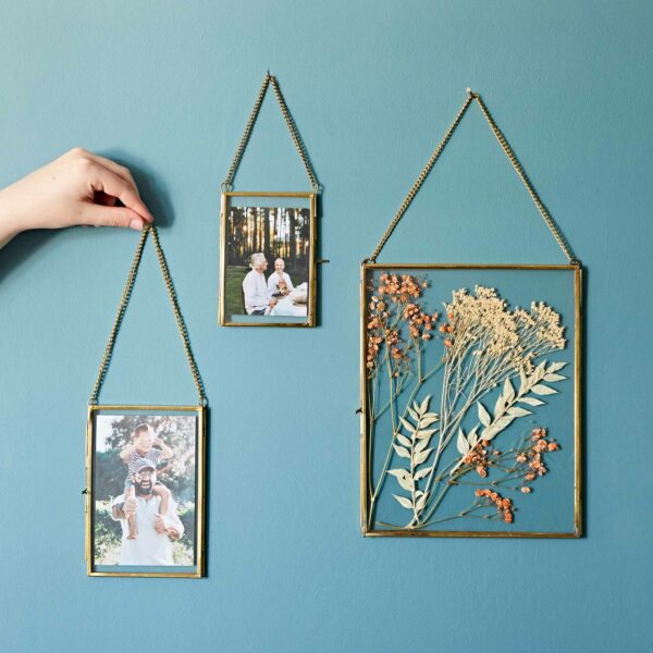 A hand adjusts one of three Dia Gold Recycled Glass Hanging Photo Frames on a blue wall. The frames, including a lovely recycled glass photo frame, contain two family photos and one dried flower arrangement.