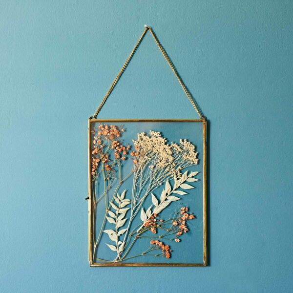 A Dia Gold Recycled Glass Hanging Photo Frame displays pressed flowers, including gold and white blooms and green leaves, all elegantly arranged on a chain against a teal wall.