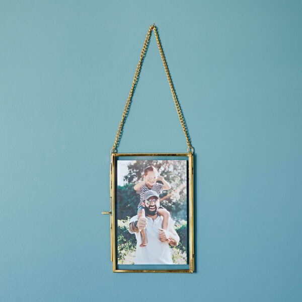 A framed photo of a man carrying a child on his shoulders is hanging on a light blue wall with a Dia Gold Recycled Glass Hanging Photo Frame.