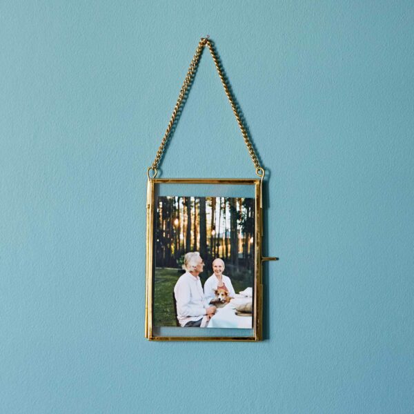 A framed photograph of two elderly people smiling and sitting outdoors, elegantly hung on a blue wall with a gold chain in a Dia Gold Recycled Glass Hanging Photo Frame.